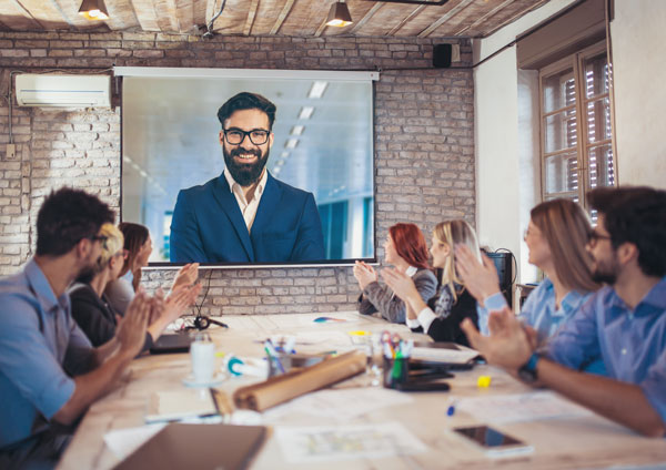 Team in einem modernen Büro applaudiert einem Redner auf einer Videokonferenz-Leinwand.